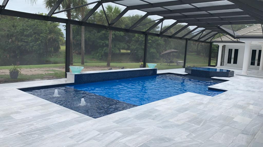 Blue swimming pool with fountains and a pale stone deck inside a screened enclosure